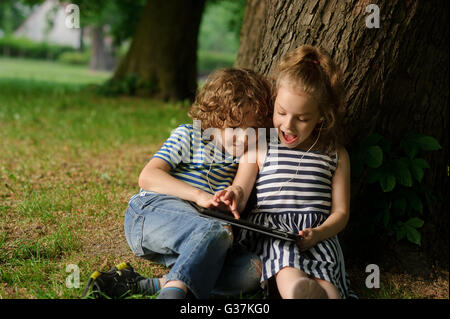 Fratello con la sorella di 7-9 anni sedersi sotto un albero e guardare nel laptop.Bambini sedersi avendo appoggiato contro un tronco di un vecchio tre Foto Stock