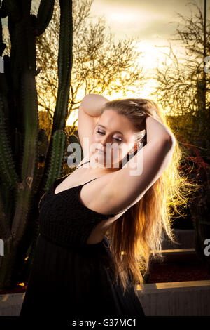 Una bella ragazza con lunghi capelli biondi pone di fronte un tramonto nel deserto. Foto Stock