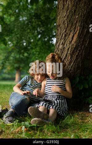 Il ragazzo e ragazza di 7-8 anni sedersi sotto un albero avente accoccolato e giocare sulla tavoletta. I bambini sono molto interessati. Essi con inre Foto Stock