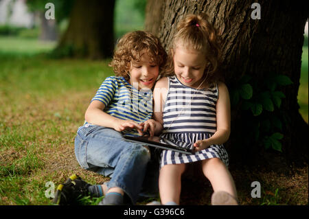 Il fratello e la sorella di 8-9 anni gioca con passione sulla tavoletta. Essi siedono avendo appoggiato contro un tronco di un grande albero. 0 Foto Stock