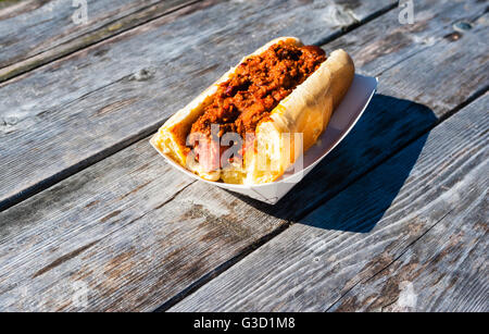 In parte mangiato salsiccia coperta in hot chili su un panino in un vassoio della carta, su weathered grigio Tavolo picnic. Foto Stock