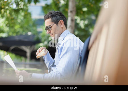 Imprenditore aziendale di mangiare il pranzo e la lettura di documenti cartacei su una panchina nel parco Foto Stock