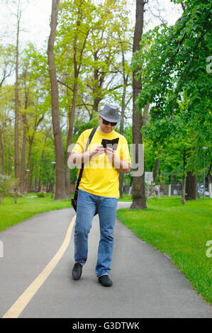 Felice l'uomo con il cappello giallo, T-shirt e il telefono è camminare nel parco Foto Stock