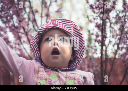 Ammirando simpatico baby girl in abiti rosa camminando tra alberi di fioritura in giardino con sorpresa di fronte Foto Stock