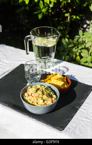 Home reso guacamole nel recipiente di ceramica e tortilla chips e crema ammuffito sul lato con la brocca e bicchiere di acqua ghiacciata. Su ardesia m Foto Stock