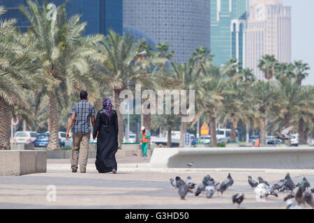 Uomo e donna che cammina su Al Corniche, West Bay, a Doha. Donna in Abaya. Lavoratore in background. Foto Stock