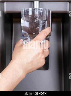 Mano azienda bicchiere mentre viene riempito con acqua filtrata dal frigorifero Foto Stock