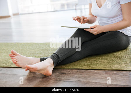 Bella giovane donna seduta sul materassino yoga amd utilizzando tablet in studio Foto Stock