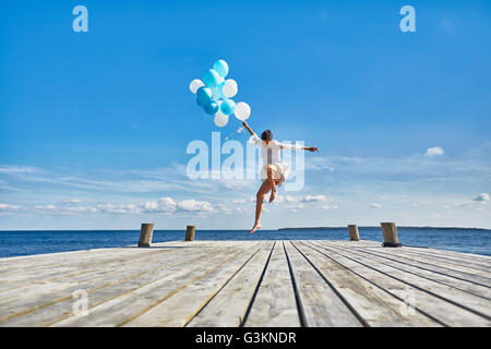 Giovane donna che danzano sul molo in legno, tenendo mazzetto di palloncini Foto Stock