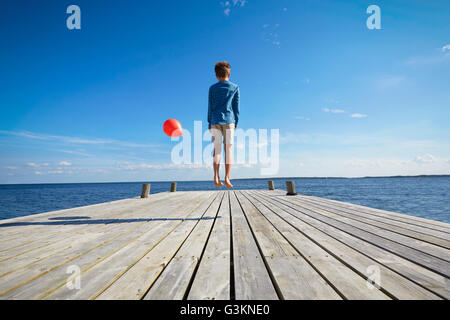 Ragazzo di saltare sul molo in legno, tenendo rosso palloncino elio, vista posteriore Foto Stock