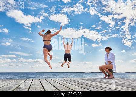 Giovane donna seduta sul post sul molo in legno, guardare gli amici salto nel mare, vista posteriore Foto Stock
