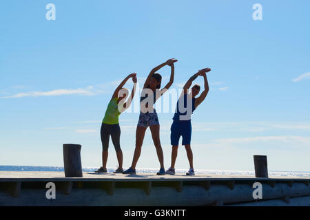 Gli amici sul molo di indossare abiti di esercizio a braccia alzate, piegarsi lateralmente stretching Foto Stock