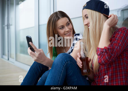 Due amiche, seduti all'aperto e di bere la birra in bottiglia, guardando a smartphone Foto Stock