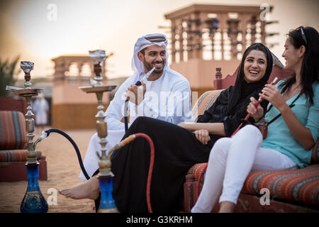 Locale giovane indossando abiti tradizionali di fumare shisha sul divano con turista femminile, Dubai, Emirati Arabi Uniti Foto Stock