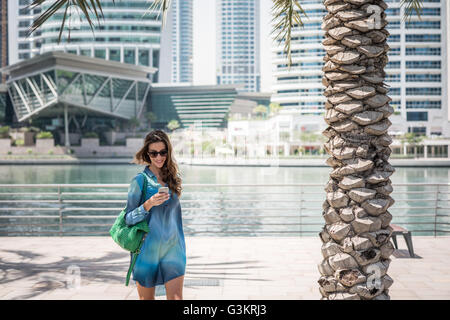 Turista femminile passeggiando sul lungomare di lettura di testi di smartphone, Dubai, Emirati Arabi Uniti Foto Stock