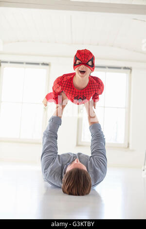 Padre giacente sul piano tenendo premuto fino figlio indossando il costume del supereroe Foto Stock