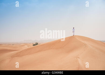 I giovani del medio oriente uomo che indossa gli abiti tradizionali guardando dalle dune del deserto, Dubai, Emirati Arabi Uniti Foto Stock