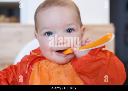 8 mese baby boy avente il suo cereale poltiglia. Indossa un cartellino arancione. Cercando di mangiare in modo indipendente Foto Stock