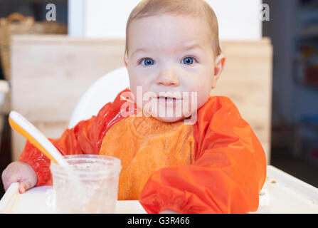 8 mese baby boy avente il suo cereale poltiglia. Indossa un cartellino arancione. Cercando di mangiare in modo indipendente Foto Stock