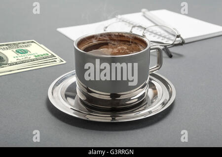 Still Life foto con tazza di caffè, prenota, penna, denaro e occhiali su sfondo grigio. Il fuoco selettivo sulla tazza di caffè. Foto Stock