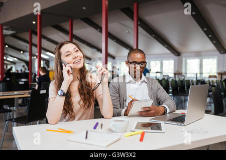 Sorridente giovane imprenditrice parlando al telefono cellulare mentre il suo collega sta lavorando in ufficio Foto Stock