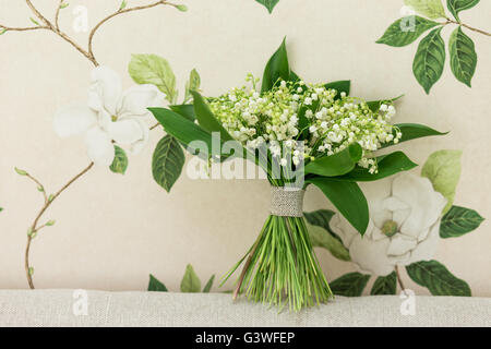 Piccolo bouquet di giglio della valle dei fiori in piedi sul tessuto decorativo con sfondo floreale nel retro. Decorazioni floreali idea. Foto Stock