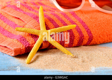 Primo piano di una stella gialla su un mucchio di sabbia e un paio di occhiali da sole su un arancio telo da spiaggia, posto su un rustico woode blu Foto Stock