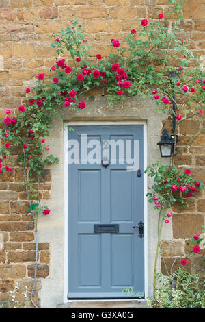 Rose rosse attorno a un legno grigio cottage in pietra porta. Adderbury, Banbury, Oxfordshire, Inghilterra Foto Stock