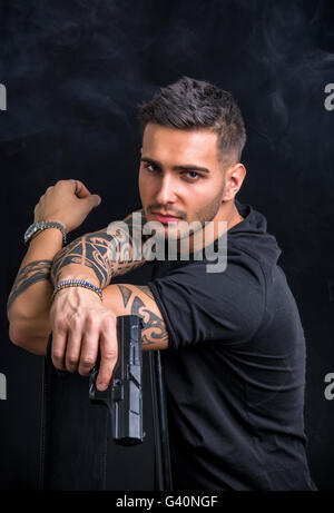 Giovane uomo bello tenendo una mano pistola, indossando t-shirt nera su sfondo scuro in studio Foto Stock