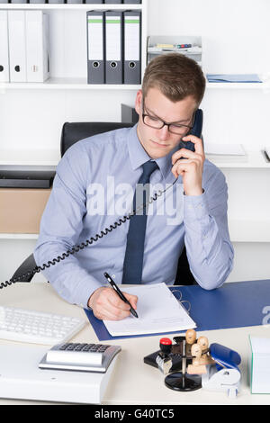 Giovane imprenditore con occhiali è di telefonare e di rendere note in un file mentre seduto alla scrivania in ufficio. Un ripiano è in Foto Stock