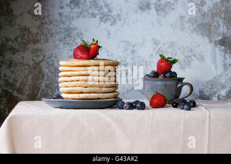 La prima colazione con pancake con fragole fresche e i mirtilli in blu piastra in ceramica e in tazza piccola, servita sulla biancheria da letto bianca ta Foto Stock