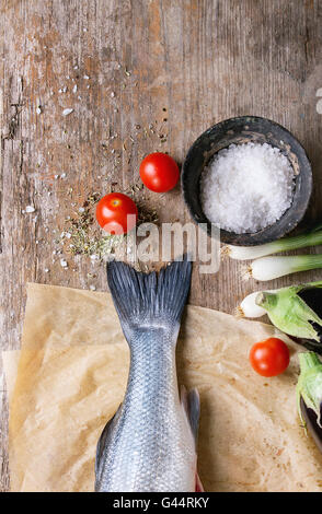 Raw seebass crudo pesce sulla carta da forno con sale marino, secca erbe e verdure su un vecchio sfondo di legno. Vista superiore Foto Stock