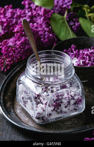 Square vasetto in vetro di lilla fiori in zucchero, cucchiaio d'argento e il ramo di Lilla sul vassoio vintage nero su sfondo di legno. Lay piatto Foto Stock