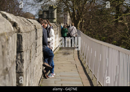 Persone che camminano lungo una strada asfaltata e si siedono su bastioni storiche delle antiche mura medievali della città vicino a Monk Bar - York, North Yorkshire, Inghilterra, Regno Unito. Foto Stock