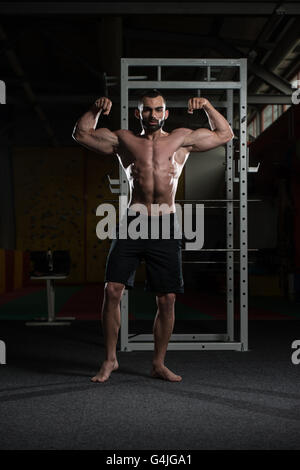 Giovane uomo in piedi forti in palestra e flessione anteriore doppia bicipite pongono - Atletico muscolare Bodybuilder Modello di Fitness in posa e Foto Stock