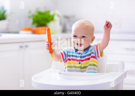 Happy Baby sitting in alta sedia mangiando la carota in una cucina bianca. Una sana alimentazione per i bambini. Foto Stock