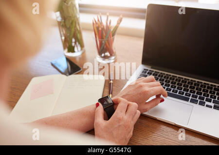 Immagine ravvicinata di donna utilizzando smartwatch. Concentrarsi sulle mani femminili e smart guarda con laptop e diario sul tavolo. Foto Stock