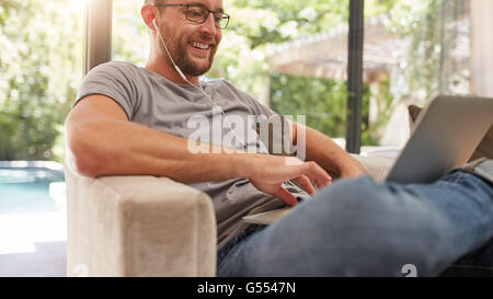 Sorridente uomo maturo usando il suo computer portatile a casa, egli è seduto sul divano con auricolari. Foto Stock