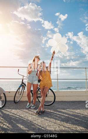 Colpo all'aperto di due donne in piedi insieme con le biciclette e palloncini sul lungomare. Migliori amici divertendosi sul loro "vacatio Foto Stock