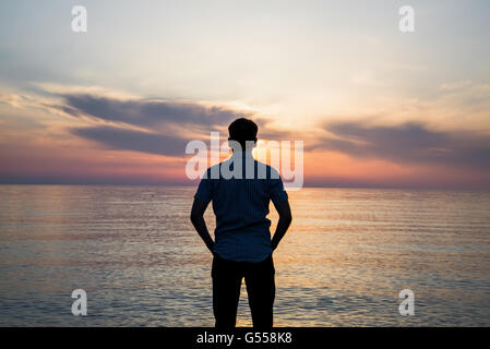 Giovane uomo in piedi presso la spiaggia di fronte all incredibile vista mare al tramonto o l'alba e il pensiero circa il suo futuro. Vista posteriore Foto Stock