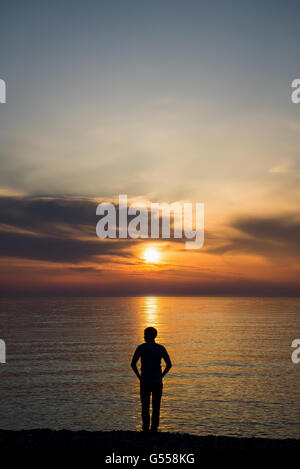 Giovane uomo in piedi presso la spiaggia di fronte all incredibile vista mare al tramonto o l'alba e il pensiero circa il suo futuro. Vista posteriore Foto Stock