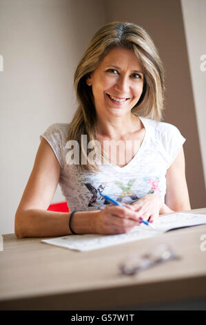 Felice donna di mezza età completando cruciverba come si siede a un tavolo a casa grinning alla fotocamera, vicino la parte superiore del corpo Foto Stock
