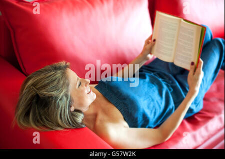 Attraente donna matura sorridente in godimento come lei legge un libro mentre un momento di relax a casa su un rosso di divano in pelle Foto Stock