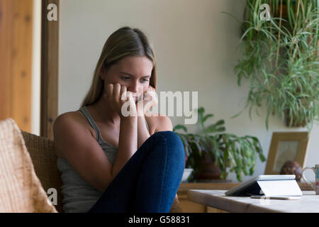 La donna a casa alla ricerca ha sottolineato nella sua sala da pranzo. Ella ha una tavoletta digitale e uno smartphone di fronte a lei. Foto Stock