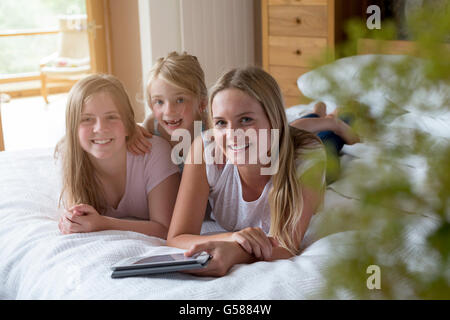 Sorelle giacente su un letto nella loro casa con una tavoletta digitale, smmiling per la fotocamera. Foto Stock