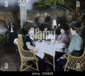 Teatime auf der RMS Titanic, Damen sitzen um einen kleinen Tisch und plaudern, dabei spielt die Bordkapelle. Il viaggio inaugurale del Titanic 1912 - Titanic disaster - Tea time a bordo per i passeggeri della prima classe - Carl Simon, lato vetro colorato slitta - storia, storico Foto Stock