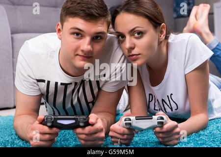 Attraente giovane coppia giacendo su un tappeto nel loro salotto per giocare con i videogiochi guardando verso la telecamera Foto Stock