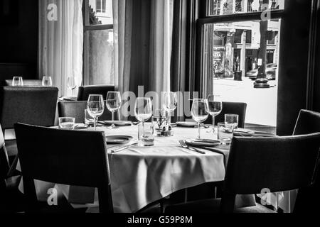 Una scarsa illuminazione di interni eleganti tavoli con tovaglie di lino in una sala da pranzo con posateria e bicchieri di vino con windows Foto Stock