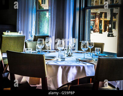 Una scarsa illuminazione di interni eleganti tavoli con tovaglie di lino in una sala da pranzo con posateria e bicchieri di vino con windows Foto Stock