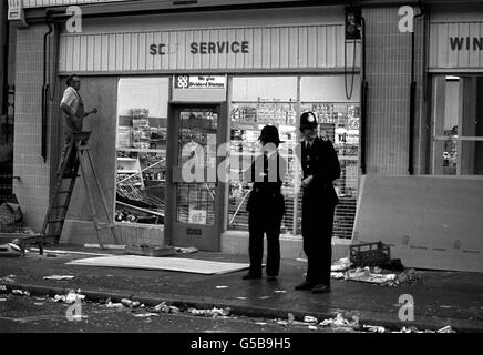 NOTTING HILL 1976: Guardia di stand della polizia come un addetto al lavoro sale le finestre del supermercato Co-op e fuori licenza a Westbourne Park Road seguendo le scene di violenza della scorsa notte a Notting Hill, Londra ovest, quando un carnevale indiano occidentale è esploso in una rivolta. Foto Stock